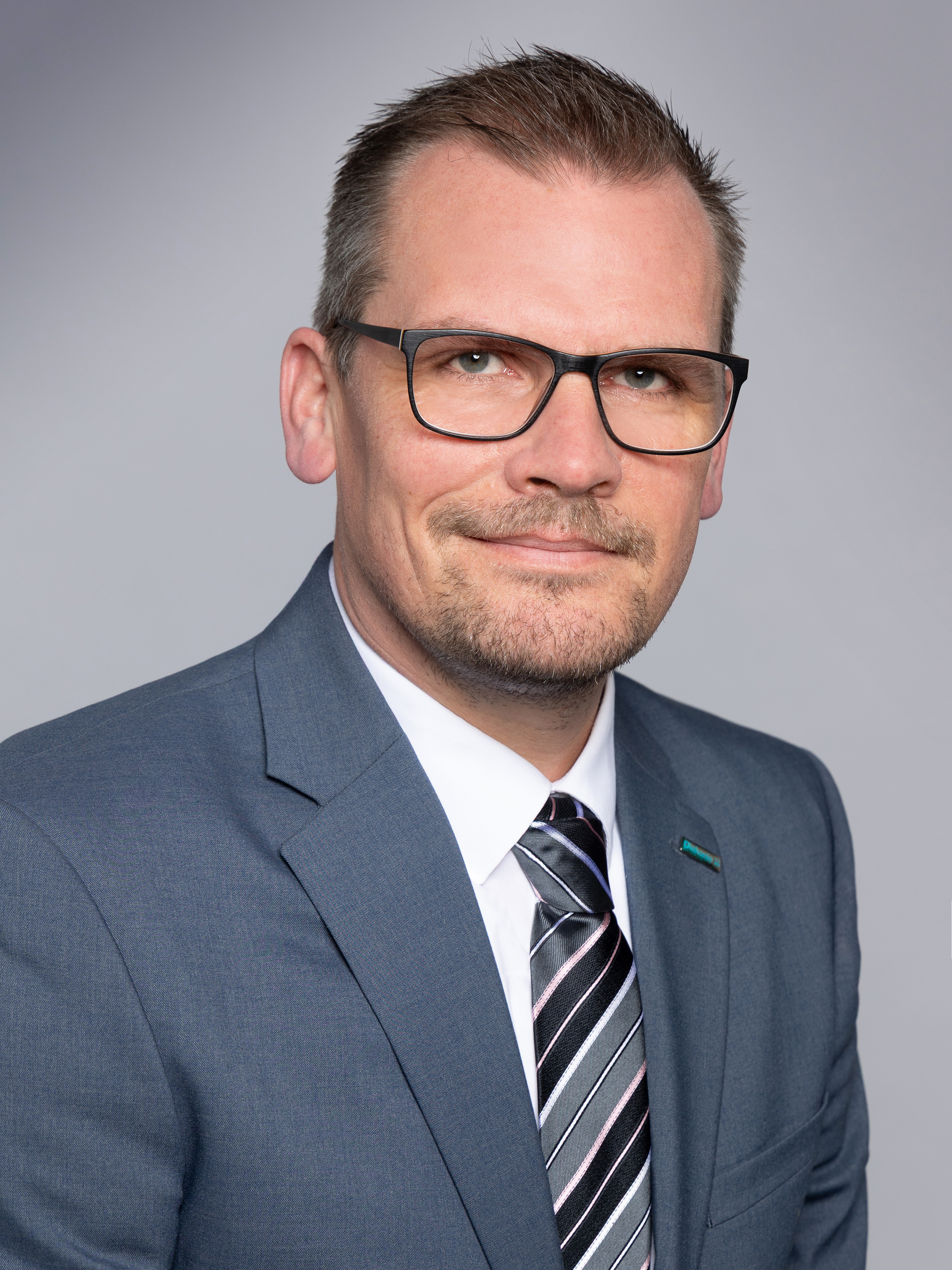 Profilfoto von Oliver Genz. Er trägt einen Anzug, trägt eine Brille. Neutraler Hintergrund.